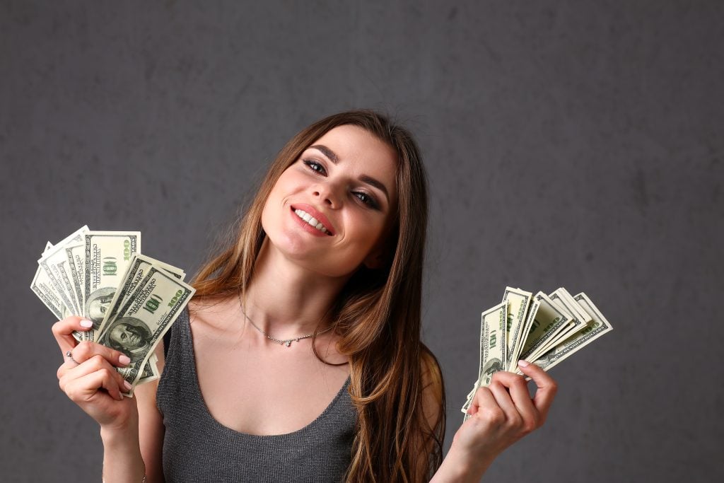 Smiling woman holding several 100-dollar bills in both hands against a dark gray background.
