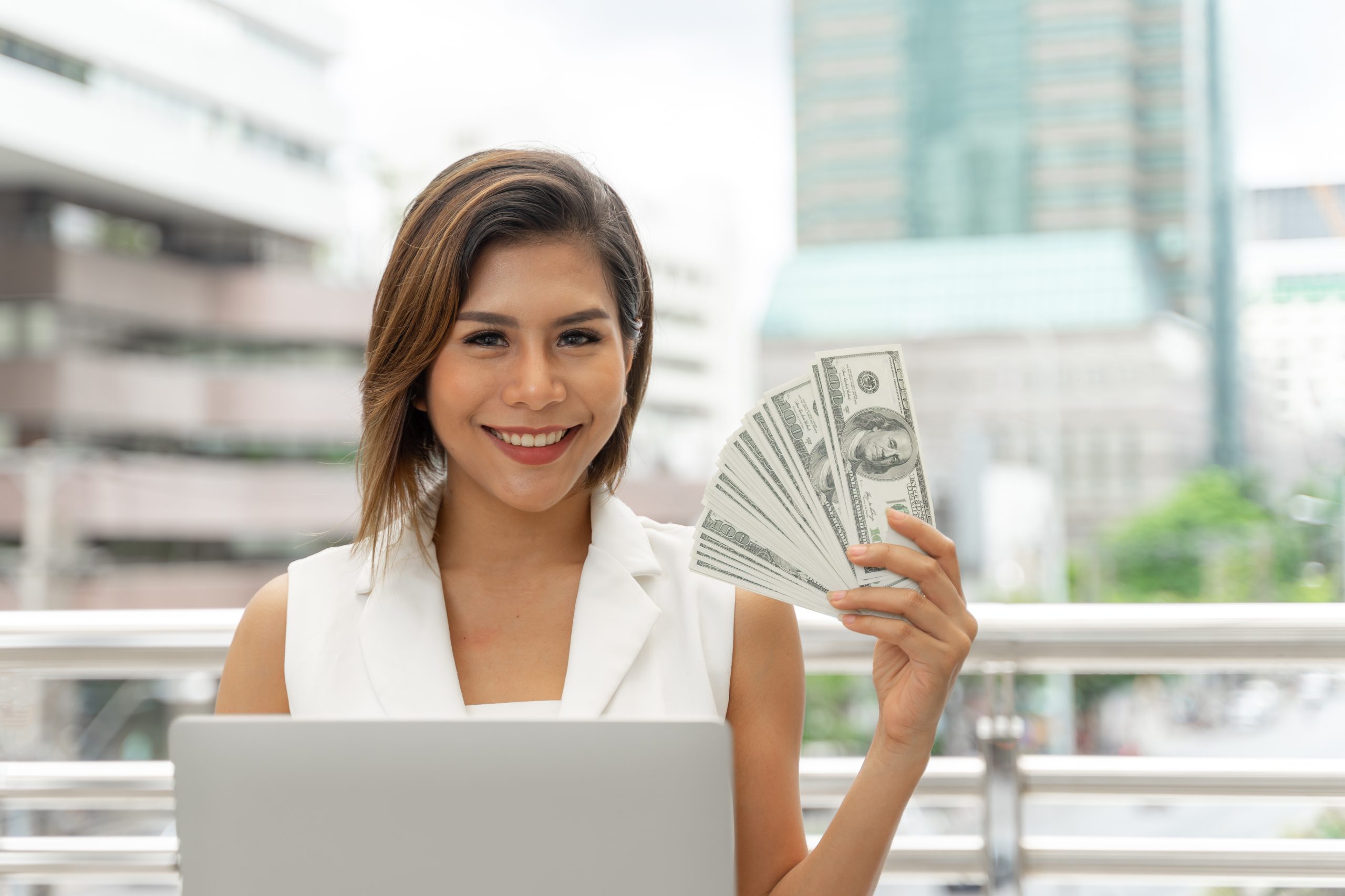 Smiling businesswoman using a laptop while holding a fan of US dollar bills in an outdoor urban setting.