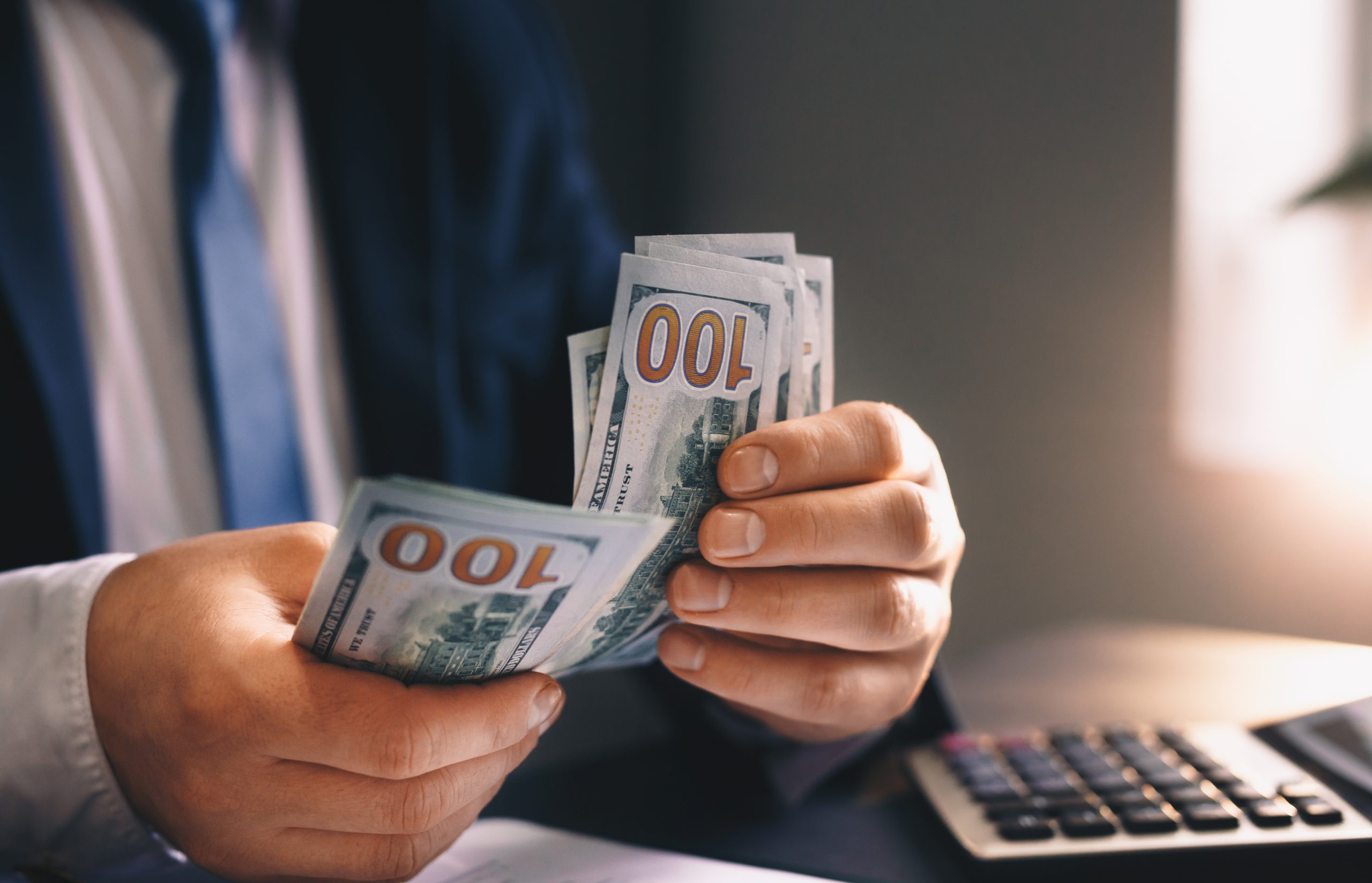 Businessman placing US dollar bills into his suit pocket against a light background.