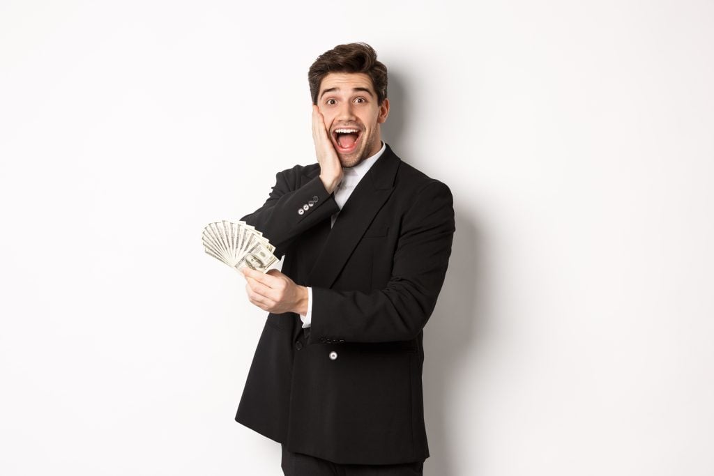 Surprised man in a black suit holding a fan of dollar bills against a light gray background.